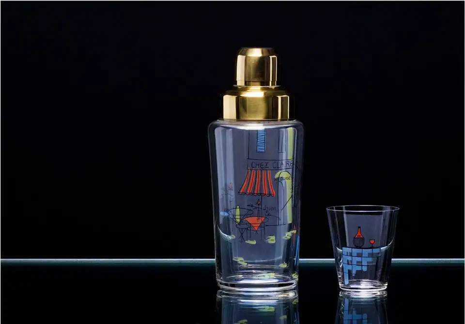 A retro glass cocktail shaker with a gold cap and a matching printed glass on a dark reflective shelf