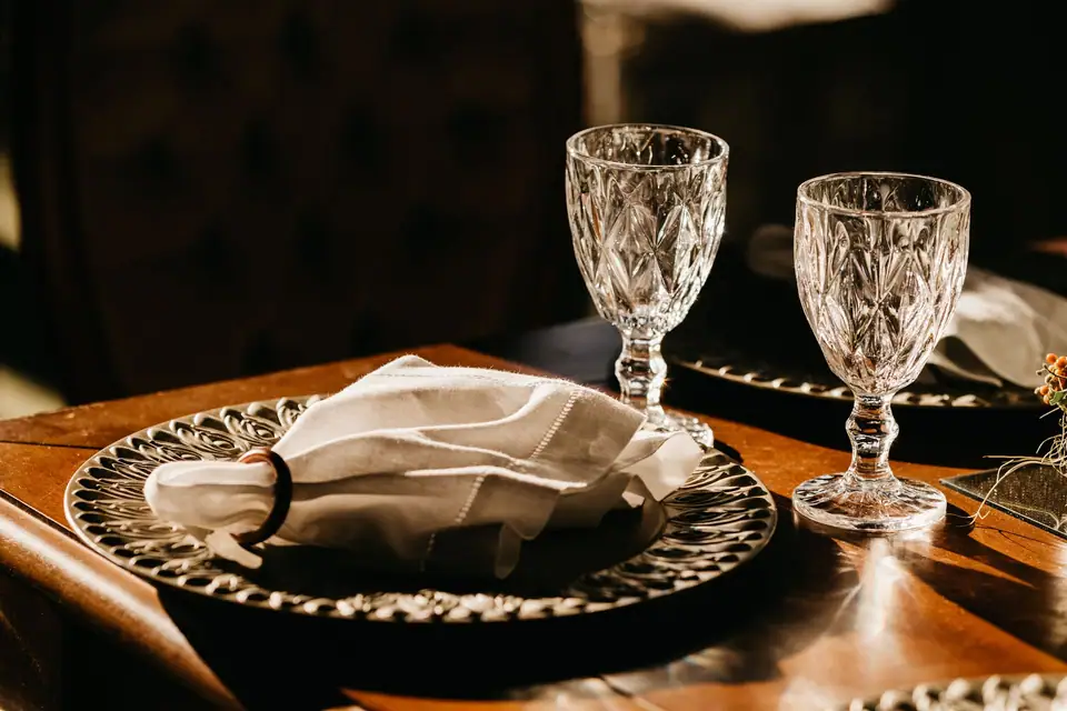 Two diamond-patterned glass goblets beside a linen napkin on a charger plate at a wooden dining table