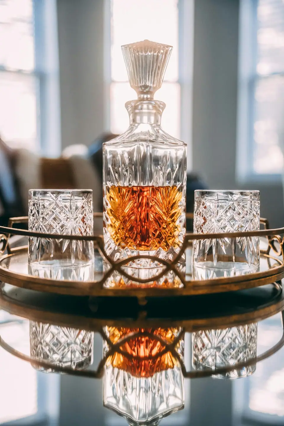 A cut crystal whisky decanter and matching tumblers on a round gilded tray in soft window light