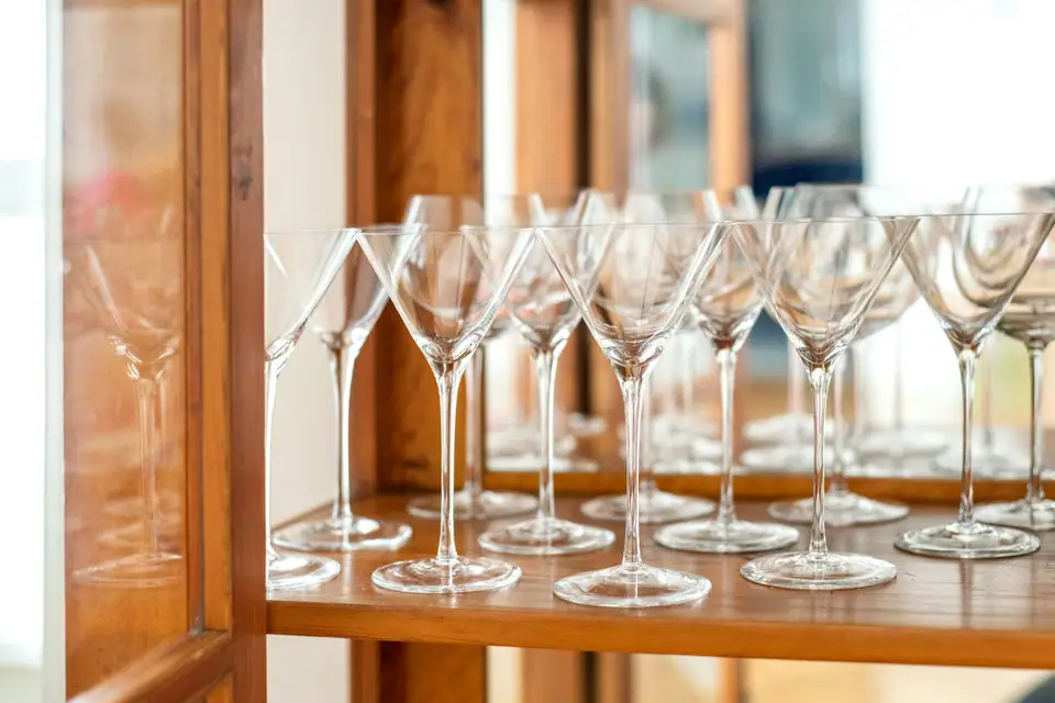 Rows of stemmed martini glasses lined up on a warm wooden bar shelf in soft daylight