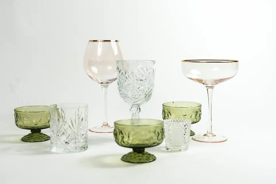 A mix of mid-century style glassware including a gold-rim coupe, cut crystal goblet and green footed dessert glasses on a white surface