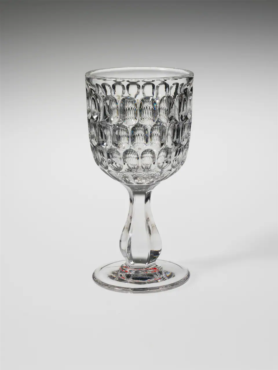 A clear pressed glass champagne goblet with a deep bowl covered in oval facets on a baluster stem, made about 1850 to 1870