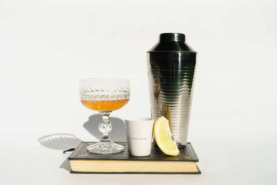 A ribbed silver cocktail shaker beside a cut crystal coupe of amber drink and a lemon slice on a vintage book, on a light background
