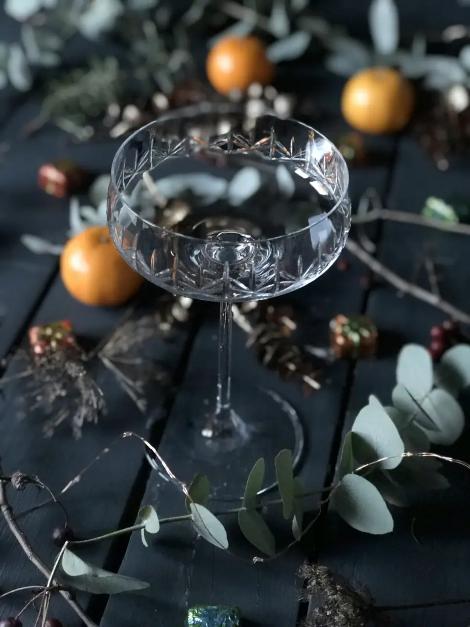 An etched crystal champagne coupe on a dark wooden table surrounded by oranges and eucalyptus sprigs
